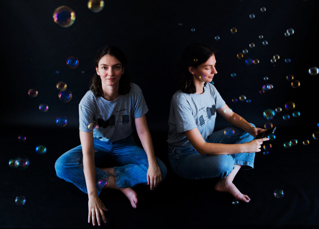 deux jeunes femmes très ressemblantes sont assises dans un décors de bulles, la première regarde le spectateur et la seconde écrist sur son téléphone.